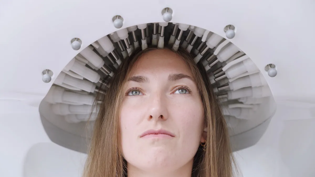 Close-up of a woman's face looking upward; the underside of a white device surrounds her head, with rows of small transducer modules reaching her hair.