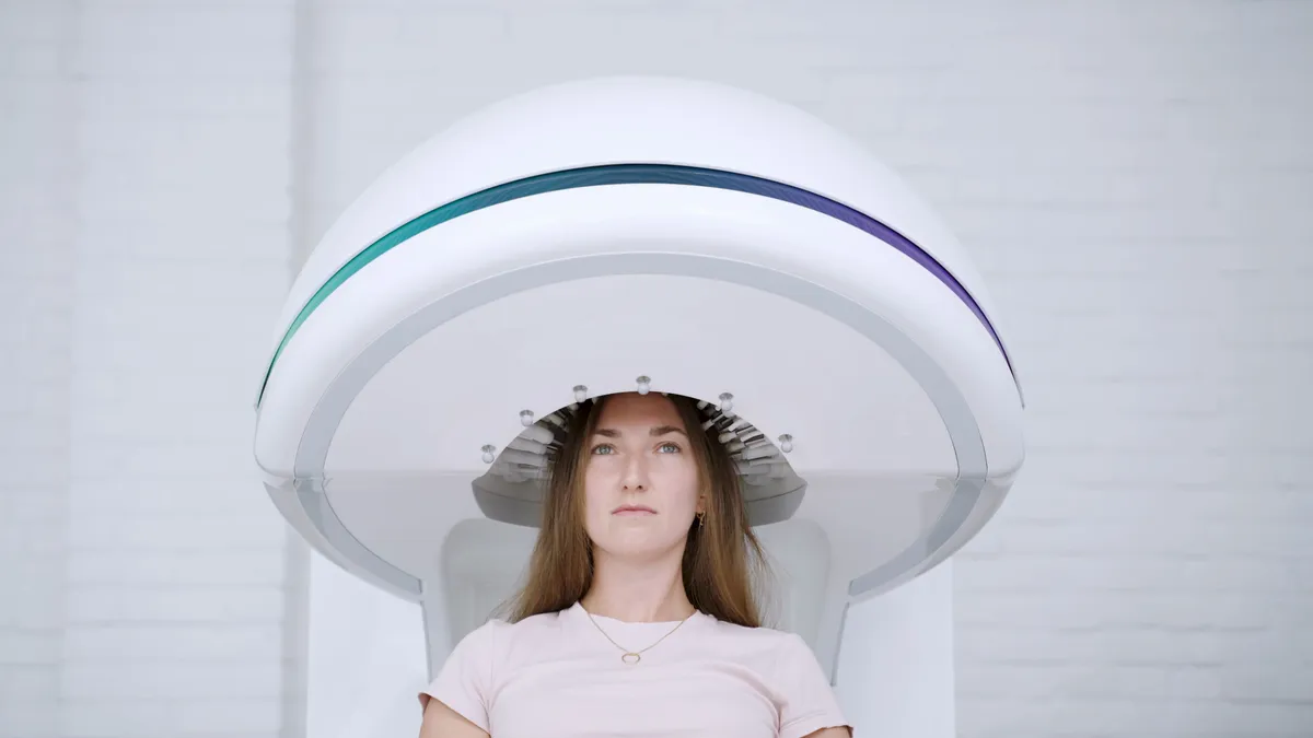 A woman sits upright in a chair, the top of her head inside a large white dome-shaped device with a green-to-purple stripe, in a bright white room.