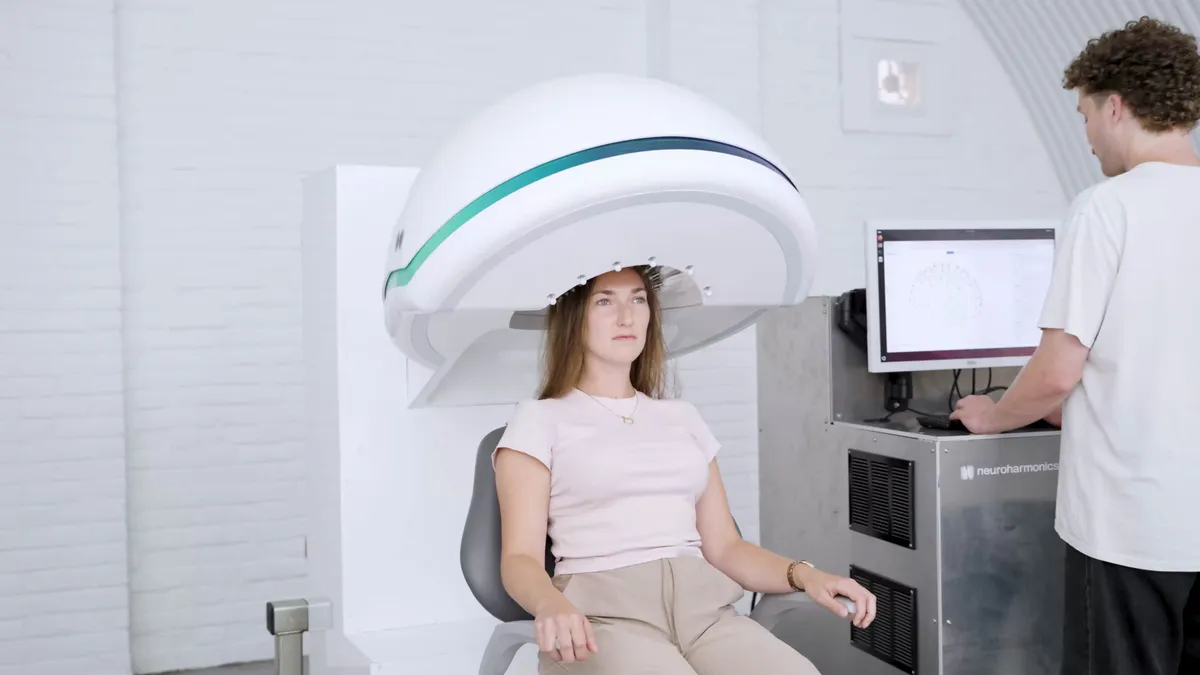 A woman seated in the white helmet-shaped device while an operator stands at a monitor displaying the system's control software.