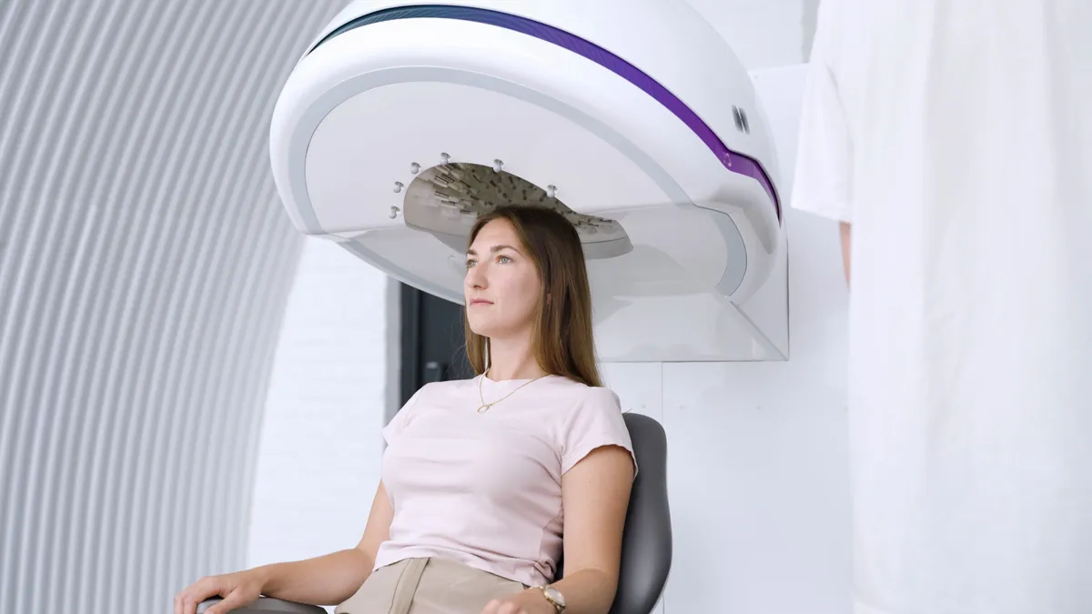 A woman sits in a chair beneath a large white dome-shaped device; the array of transducer modules is visible inside the dome above her head.