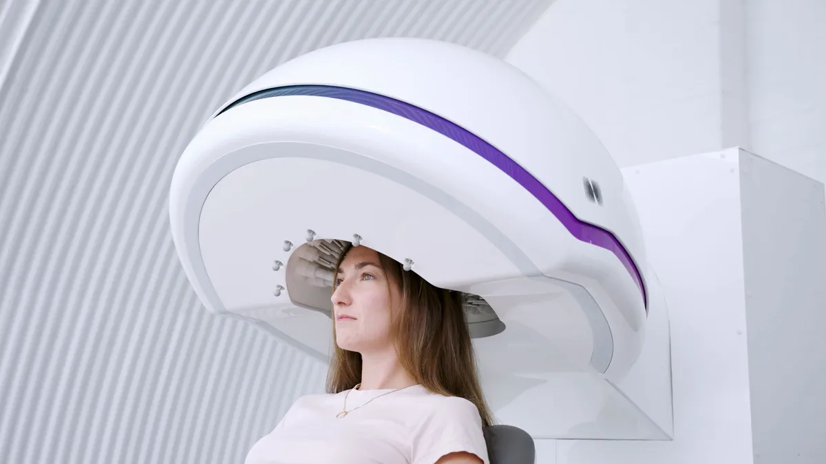 Side profile of a woman whose head is inside a large white helmet-shaped device mounted on a stand, with a purple stripe along its rim.