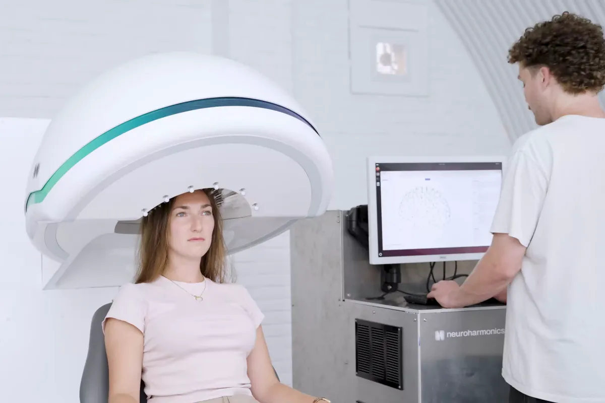 A participant with long, unshaved hair sits in a chair beneath a white dome-shaped helmet, while an operator adjusts settings on a console showing a map of the transducer array.